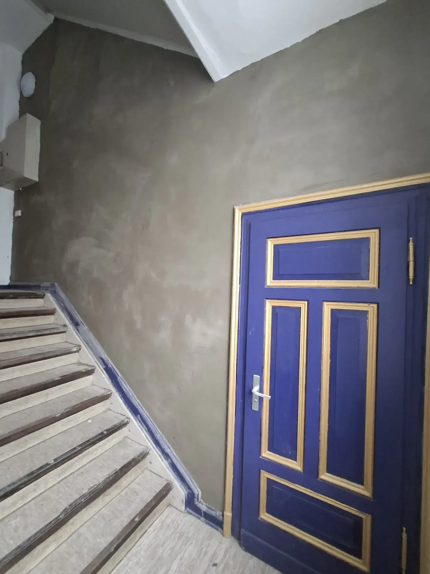 Treppenhaus vorher: roher, unbehandelter Wandputz neben der blauen Wohnungstür